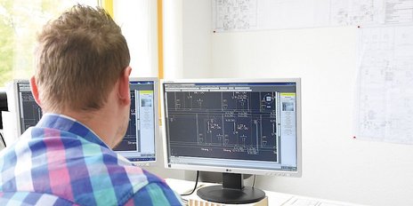 Ein Mann mit einem karierten Hemd sitzt vor zwei Monitoren, die technische Zeichnungen anzeigen, in einem hellen Büro.