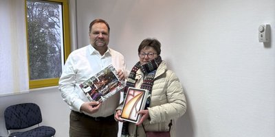 Ein Mann und eine Frau stehen in einem hellen Raum und halten jeweils ein Bild in der Hand, beide lächeln freundlich.