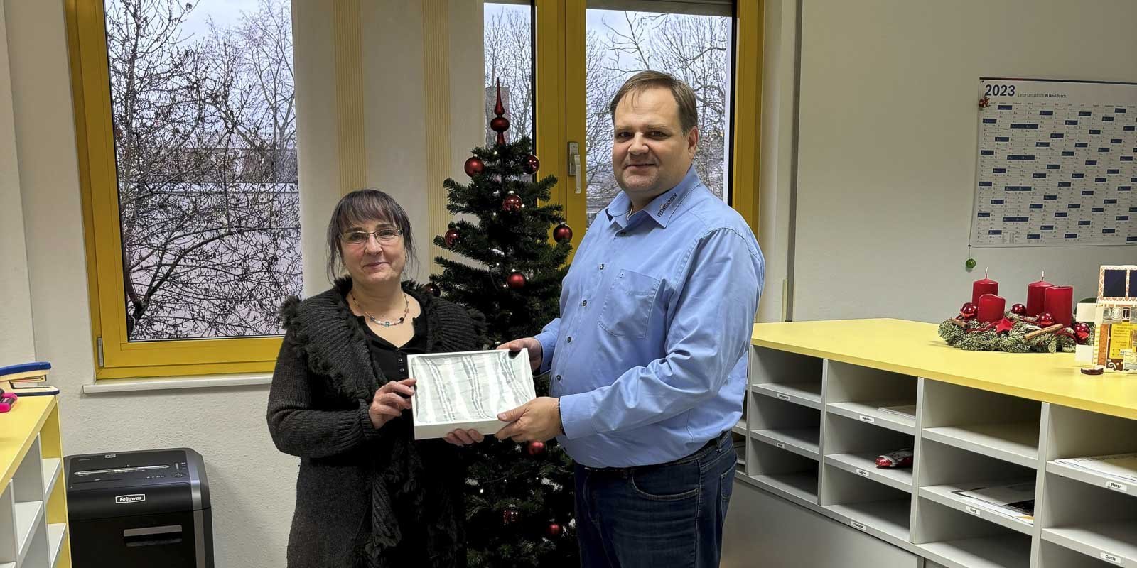 Eine Frau und ein Mann stehen in einem Büro, während sie ein Geschenk vor einem Weihnachtsbaum überreichen.