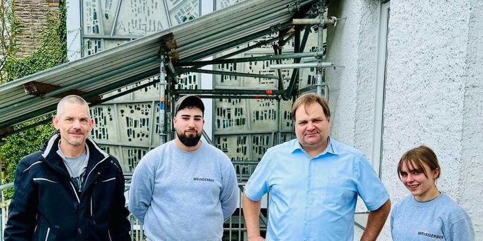 Vier Personen stehen vor einem Gebäude mit einem Gerüst, lächelnd und in Arbeitskleidung, bereit für ihre Aufgabe.