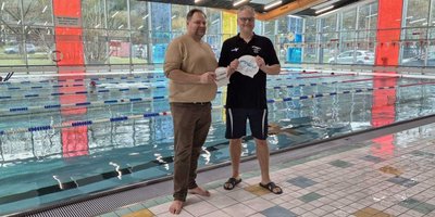 Zwei Männer stehen am Beckenrand eines Schwimmbads und halten Schwimmkappen in den Händen, lächelnd und entspannt.