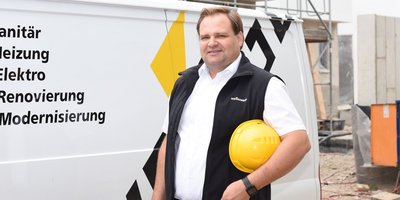 Ein Mann in Arbeitskleidung steht vor einem Lieferwagen, lächelt und hält einen gelben Helm in der Hand.